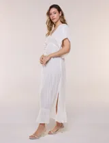 Vestido largo blanco confeccionado en viscosa con hilos de lurex, escote en V profundo con lazo y borlas, mangas kimono y falda fluida con volados en el ruedo.