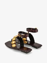 Sandalia plana de cuero sintético color marrón, con punta cuadrada y diseño de tiras anchas adornadas con apliques metálicos dorados en forma de óvalos. Cuenta con una correa ajustable al tobillo con hebilla metálica.