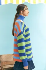 Sweater de tejido de punto grueso, cuello alto con cierre parcial y manga larga. Presenta un diseño de bloques de color y rayas horizontales en tonos vibrantes como azul, verde lima, naranja y celeste.