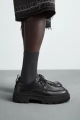 Zapato de vestir tipo derby, color negro, con detalle de bordón en la puntera. Cuenta con tirador trasero para facilitar el calce, horma redonda y suela con volumen y diseño irregular.