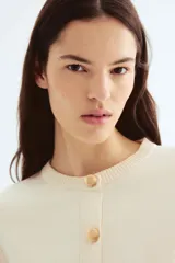 Cárdigan corto de punto fino color crema, con cuello redondo, cierre frontal con botones dorados y bolsillos delanteros decorativos con botón.