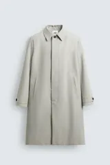 Trench coat de corte relajado, color gris claro, confeccionado en tejido técnico con mezcla de algodón. Presenta cuello solapa, manga larga con puño ajustable, bolsillos de doble vivo en cadera, bolsillo interior y abertura en el centro de la espalda. Cierre frontal con botonadura oculta.