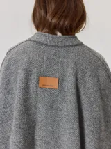 Capa de tejido de punto en color gris melange, con diseño abierto y cierre frontal mediante un botón tipo duffle de madera con detalle en cuero. Presenta bolsillos laterales amplios y bordes con costuras a la vista.