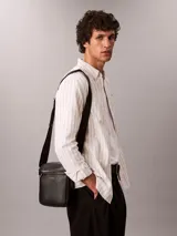 Cartera reporter estructurada Calvin Klein de poliuretano con textura granulada, color negro, cierre superior de cremallera y bolsillo frontal con cierre. Cuenta con correa de hombro ajustable y logo metálico de la marca en la parte delantera.
