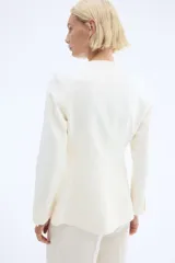 Blazer entallado color blanco roto, con cuello redondo, sin solapas, manga larga y botones frontales de madera. Posee bolsillos de parche delanteros y forro interior.