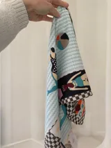 Pañuelo cuadrado de seda con estampado temático de playa, que incluye figuras de personas, pelotas de playa y patrones geométricos en tonos celeste pastel, rosa, bordó, naranja, negro y blanco.