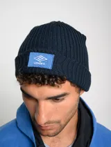 Gorro tipo beanie tejido en punto acanalado, color azul marino, con parche frontal rectangular en color celeste que presenta el logo de Umbro bordado en blanco.