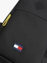 Mochila negra de poliuretano con compartimento principal y cierre de doble cremallera con tiradores amarillos a contraste. Cuenta con bolsillo frontal con cremallera, asa superior, correas ajustables y acolchadas, y parche engomado de Tommy Jeans en la parte delantera.