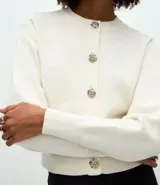 Cardigan de punto color negro, con cuello redondo, cierre frontal con botones metálicos en forma de flor y mangas largas con puños acanalados.