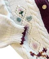 Cardigan de lana merino color marfil, con bordados florales artesanales, trenzas y rombos calados en el frente. Posee cierre frontal con botones vintage dorados y terminaciones en crochet celeste en cuello, puños, ruedo y tira de botones.