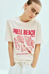 Remera de algodón de corte holgado en color crema, con cuello redondo y mangas cortas. Presenta un estampado frontal en color rojo con ilustraciones de caracoles de mar y el texto 'SHELL BEACH Club'.