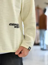 Sweater de punto liviano color beige, con textura marcada, cuello redondo y mangas largas de corte relajado, con pequeño estampado tipográfico negro en el lateral inferior.