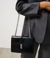Cartera tipo shoulder bag de tamaño mediano, confeccionada en poliuretano con textura efecto croco. Presenta una tapa frontal con cierre metálico de giro, asa de mano reforzada y una correa larga de cadena plateada. Diseño estructurado con acabado sofisticado.