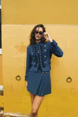 Blazer corto de tweed en color azul, con cuello mao y manga larga. Presenta detalles de pasamanería negra en el frente y cierre con ganchos metálicos y falsos botones dorados.