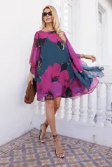 Vestido corto de corte amplio y fluido, con mangas tipo capa y escote redondo. Presenta un estampado floral de grandes flores fucsias sobre un fondo azul oscuro.