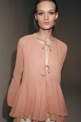 Blusa de viscosa color rosado con cuello redondo, manga larga abullonada con puños elásticos y cierre frontal con lazadas. Presenta un diseño con volantes en el bajo y corte asimétrico.