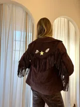 Campera marrón de gamuza con flecos en la espalda y las mangas, solapas anchas y detalles de pines metálicos en forma de estrella y figuras abstractas.