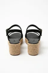 Sandalia de gamuza color negro con plataforma de yute trenzado, tira ancha en la parte delantera y tira ajustable al tobillo con hebilla metálica dorada.