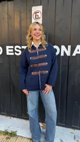 Campera de estilo militar en color azul marino, confeccionada en tejido de punto grueso. Presenta un diseño de cuello redondo, cierre frontal con tiras de cuero marrón y botones metálicos decorativos. Posee un corte recto y holgado.
