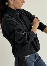 Campera de jean azul oscuro, estilo bomber, con cuello camisero y cierre frontal. Presenta un parche rectangular bordado en la espalda con patrón de rombos y texto.