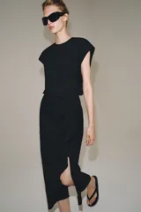 Vestido midi negro con cuello redondo y manga sisa, confeccionado por Zara Woman Collection. Presenta un diseño drapeado en la parte delantera y un bajo asimétrico con un voluminoso volante lateral. Incluye un pequeño cierre de botón en la espalda.