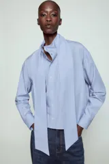 Camisa de algodón color celeste, con cuello alto y lazo integrado en el mismo tejido. Presenta cierre frontal con botones, manga larga con puños y un bolsillo frontal en el pecho. Diseño de corte recto.