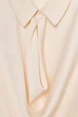 Camisa corta de manga larga, color crema o beige muy claro, con cuello solapa y escote drapeado o fluido tipo cascada.