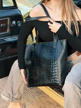 Bolso tipo shopper negro con textura símil cocodrilo, doble asa de hombro y dije colgante con forma de moño.