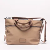 Bolso tipo shopper/bandolera color beige/caqui, con dos asas cortas de tela y una correa larga ajustable y desmontable. Presenta un bolsillo horizontal con cierre en la parte frontal y un parche rectangular de material liso en la parte inferior central.
