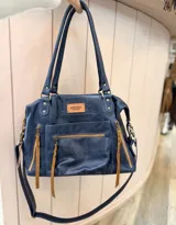 Cartera tipo tote bag de cuero color negro, con múltiples bolsillos externos con cierres metálicos y tiras decorativas en tono tostado. Cuenta con doble asa de hombro y correa larga ajustable y desmontable.