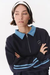 Buzo estilo rugby de corte oversized, color verde oscuro, con cuello tipo camisa en denim azul claro y cierre frontal parcial. Presenta un recorte horizontal en el pecho, logo bordado y tiras laterales en las mangas en contraste.