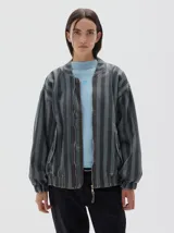 Campera bomber de denim con estampado de rayas verticales en tonos grises, cierre metálico frontal y bolsillos. Presenta un corte oversized y puños y cintura elásticos.