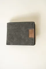 Billetera de hombre con diseño bi-fold, confeccionada en material sintético con textura. Presenta un color gris oscuro con un detalle rectangular en color marrón y costuras decorativas en contraste de color blanco.