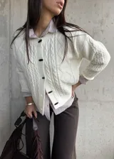 Cardigan de punto trenzado color natural, con cuello redondo, cierre frontal de botones negros y mangas largas con puños acanalados.