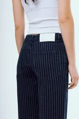 Pantalón de jean azul oscuro de corte recto y tiro medio, con diseño de rayas diplomáticas blancas. Presenta bolsillos delanteros y traseros, y cierre frontal con botón y cremallera.