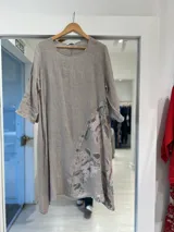 Vestido midi color beige, de corte holgado y silueta acampanada, con cuello redondo y mangas tres cuartos. Presenta un diseño cruzado asimétrico en la parte inferior con detalle de botones y estampado floral en tonos rosados y verdes.