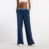 Pantalón deportivo ancho de mujer New Balance modelo Track Pant, color azul marino. Presenta cintura elástica con ribete de rayas blancas y un pequeño logo bordado en el muslo. Tiene aberturas laterales en el ruedo.