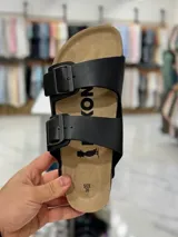 Sandalias de dos tiras negras con hebillas metálicas ajustables, plantilla anatómica y suela plana.