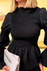 Blusa negra de manga larga con diseño de nido de abeja en el pecho y la cintura, cuello alto y mangas abullonadas con puños ajustados.