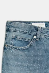 Jeans de corte recto y tiro bajo con cinco bolsillos, trabillas para cinturón y cierre frontal con cremallera y botón metálico. Presenta un acabado con aplicación de brillos en la parte delantera.