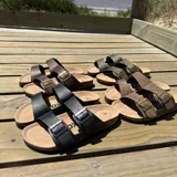Sandalia tipo Birkenstock con doble tira ajustable de cuero sintético negro y hebillas metálicas. Cuenta con plantilla anatómica de corcho y suela de goma.