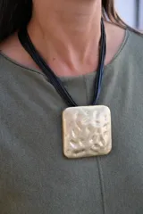 Collar de diseño con dije cuadrado de gran tamaño en tono dorado con textura irregular y martillada, suspendido de múltiples cordones negros finos.