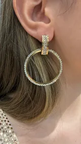 Caravanas de diseño circular en tono dorado, adornadas con incrustaciones de strass brillante en toda la estructura y en el soporte superior.