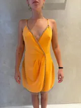 Vestido corto cruzado de color naranja con escote en V profundo, tirantes finos regulables y detalles de flecos plateados brillantes a lo largo del escote, los bordes y el dobladillo inferior. Presenta drapeado suave en la zona frontal de la falda.