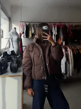 Campera puffer corta de color marrón, con diseño acolchado, cuello alto, cierre frontal con cremallera y botones a presión, y puños elásticos.