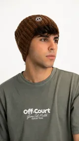 Gorro tipo beanie de punto grueso en color marrón, con textura acanalada y parche circular con logo en la parte frontal.