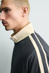 Sudadera de corte relajado en color azul marino con cuello alto y cierre frontal de cremallera. Presenta bandas laterales en color crema a contraste que recorren las mangas, puños y bajo en tejido rib, y bolsillos laterales de vivo.