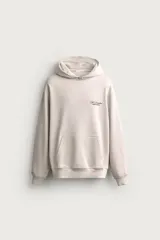 Sudadera hoodie relaxed fit de color beige, con cuello con capucha, manga larga y bolsillo frontal tipo canguro. Presenta un pequeño estampado de texto en contraste en el pecho.