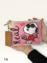Sobre de cuero color crema con diseño intervenido a mano por la artista GGIL. Presenta un estampado central en color rosa con la palabra 'real', un dibujo de Snoopy con lentes de sol y un diamante. Cuenta con cierre superior y etiqueta de cuero colgante.