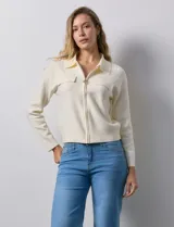 Cardigan de tejido punto color crema, con cuello tipo solapa, cierre frontal completo con tirador circular metálico y dos bolsillos de parche simulados en el frente. Presenta detalles de canalé en los hombros.